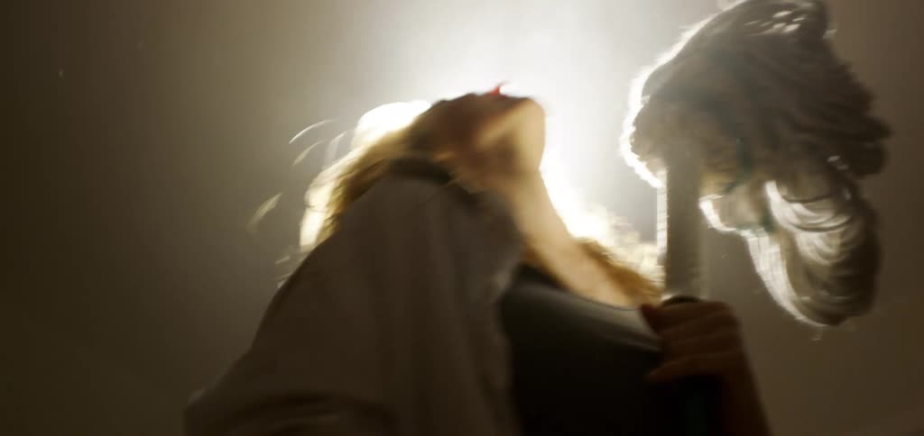 A young woman holds a mop singing and dancing with it like a dance partner while bright ceiling lights illuminate the building