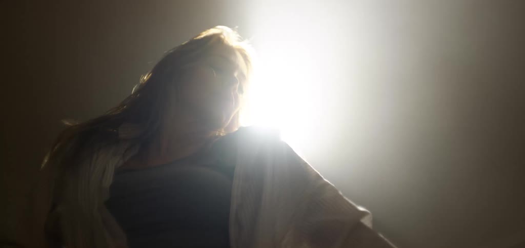 A woman with long wavy hair dances with her head back and her body swaying with a bright light shining from behind her