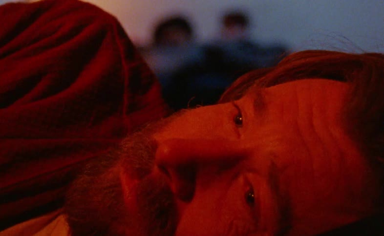 Bearded man lying down talking to two other people lying down on a bed behind him indoors