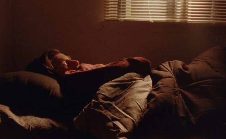 A man with light hair and a pink shirt is lying on a bed with his body covered by a dark blanket looking up and then turning his head to rest on the pillow as a shaft of light shines through the blinds onto his face