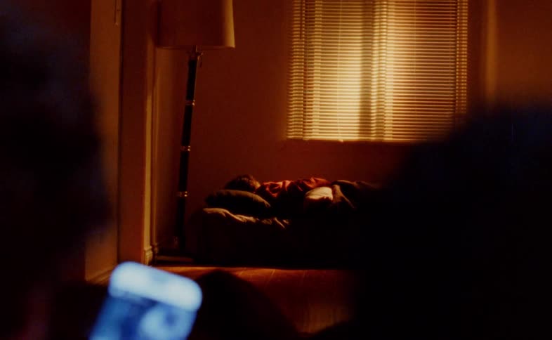 A dark bedroom with blinds on a window as a person is seen from a distance lying in bed with a second person looking down at their phone in the foreground
