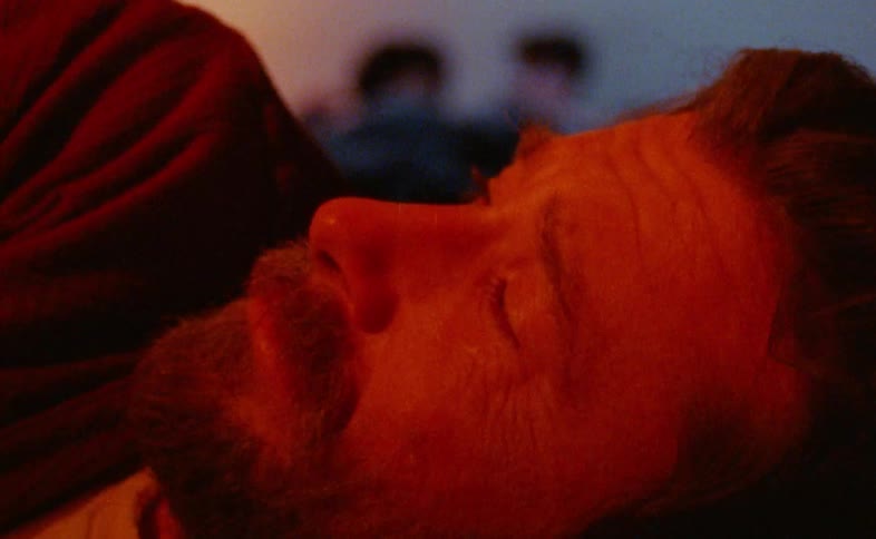 Scared and depressed bearded man looking at two other people lying down on a bed behind him indoors