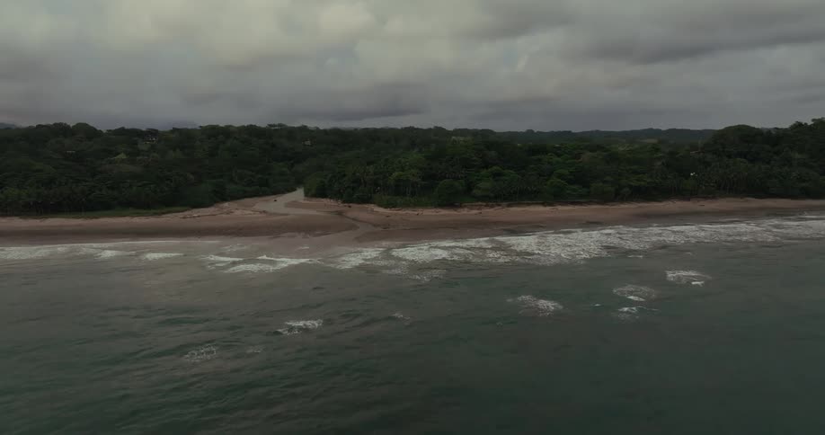 An aerial view of the ocean with waves hitting the sand on a shoreline with a jungle behind it under a cloudy sky