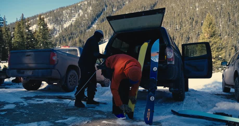 Two people wearing snowboarding outfits preparing snowboarding gear next to a vehicle with a mountain and a forest in the background outdoors during day time