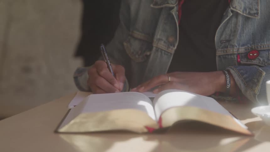 A person in a denim jacket with a spiderman patch sits at a table and writes in a notebook with a pen with a cup and a large open book also on the table