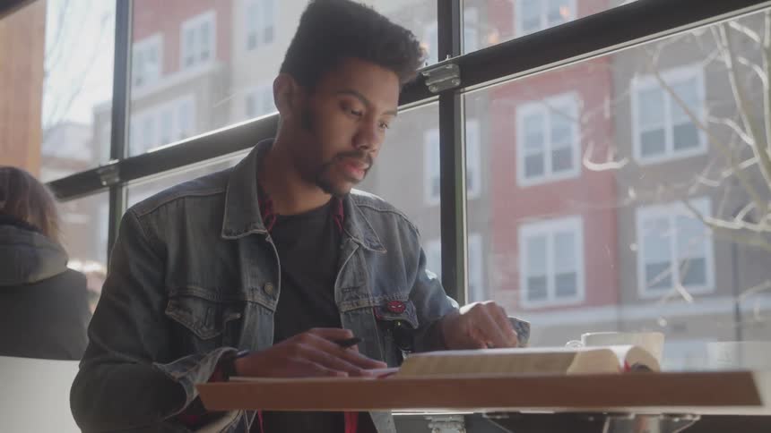 A man with a denim jacket and a black shirt sits at a table near a large window looking down at a book and taking notes