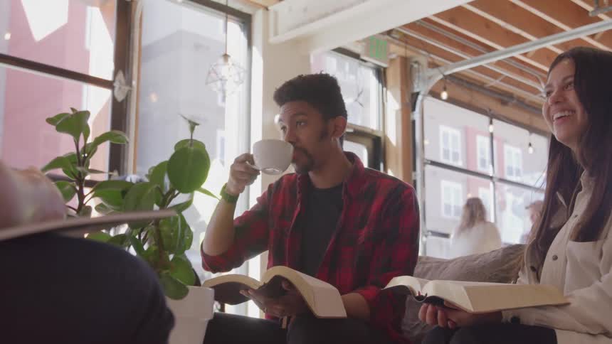 A group of people sits together some of them holding books and one person drinks from a mug