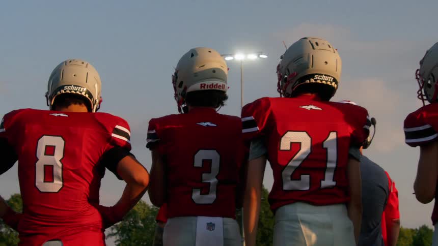 A wide shot from behind shows a row of four football players in red jerseys and white helmets with their backs to the camera they are standing on a field and looking up at the bright lights of the stadium
