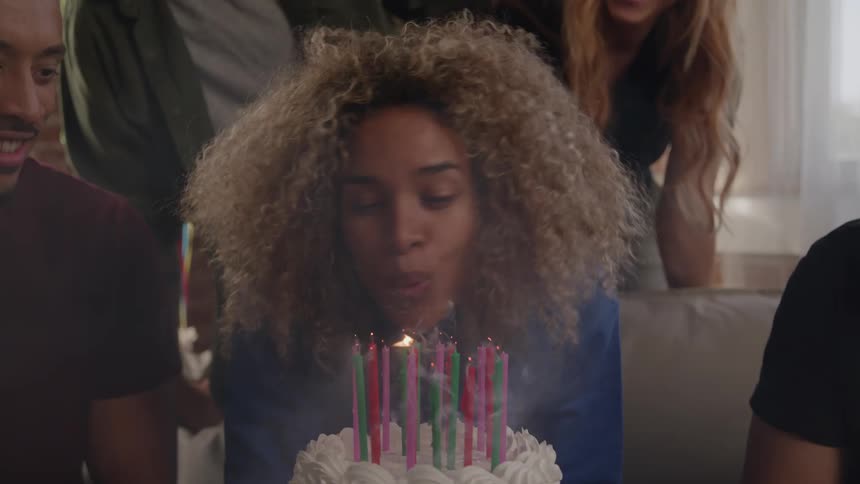 A joyful young woman with curly hair celebrates her birthday surrounded by a group of smiling friends she makes a wish and enthusiastically blows out the candles on her cake as everyone claps and cheers for her