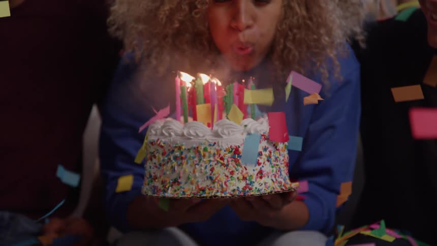 A woman sitting at a party with people around her holding a birthday cake in her hands as pieces of colored paper fall around her then she smiles and blows out the candles on the cake