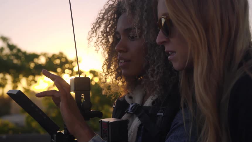 Two women stand outdoors at sunset and look at a tablet