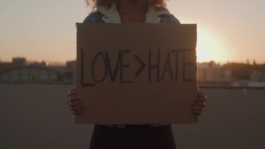 A video shows a person holding up a cardboard sign that says love hate in black letters the sign is held against the backdrop of a cityscape at sunset with the setting sun creating a strong lens flare