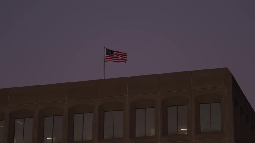 A wide shot of the top of a stone building shows an american flag flapping in the wind the sky is dark and cloudy suggesting either dawn or dusk