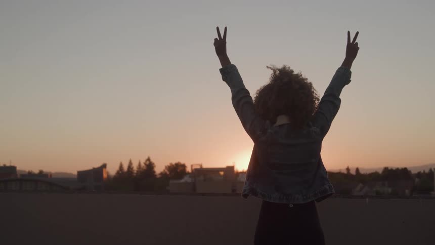 Person with arms raised making peace signs with their hands in front of the sunset