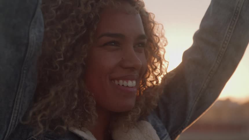 A smiling woman with long curly hair and a denim jacket stands on a rooftop with her arms raised above her head looking out at a bright sunset