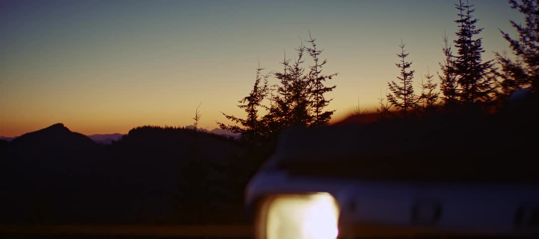 A view from a low angle shows the front of a white car or truck with its bright headlight on and a yellow and orange sunset visible behind a dark hillside with trees