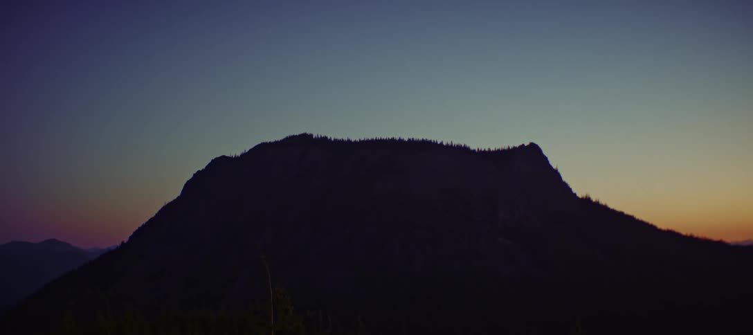 A low straight on view shows a large dark mountain with a jagged ridge of trees on top silhouetted against a colorful sunset sky that is blue at the top and transitions to orange at the bottom