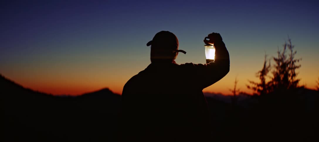 A persons silhouette is seen from behind as they hold a glowing lantern over their head and look out at a dark mountain landscape under a twilight sky