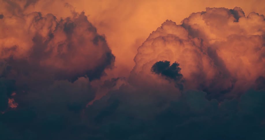 A close view shows large textured dark clouds with bright orange and pink light shining behind them as they move slowly across the frame