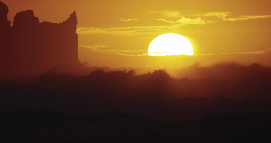 A brilliant orange sun sets over a desert landscape casting a golden hue over the clouds and sky a large dark rock formation stands tall to the left with the rest of the rocky terrain silhouetted in the foreground