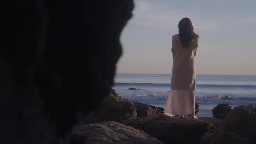 A woman stands on rocks by the ocean and watches the waves on the water and the setting sun