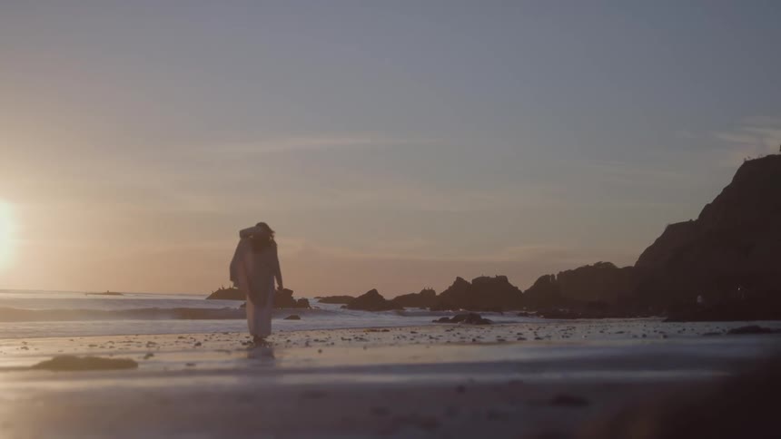 A woman in a long flowing dress stands on a wet sandy beach as waves crash on the shore behind her with a sunlit ocean and large dark rock formations on the right