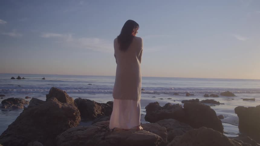 White woman standing over rocks next to the sea at the beach outdoors during dusk or dawn