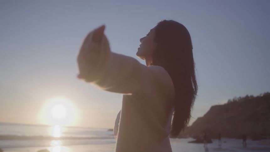 Long haired white woman standing at the beach in front of the sea and stretching her arms with other people far away on the background outdoors during dusk or dawn