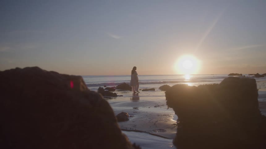 A woman in a white dress walks on a sandy beach between two large dark rocks as the sun shines and reflects on the water in the distance