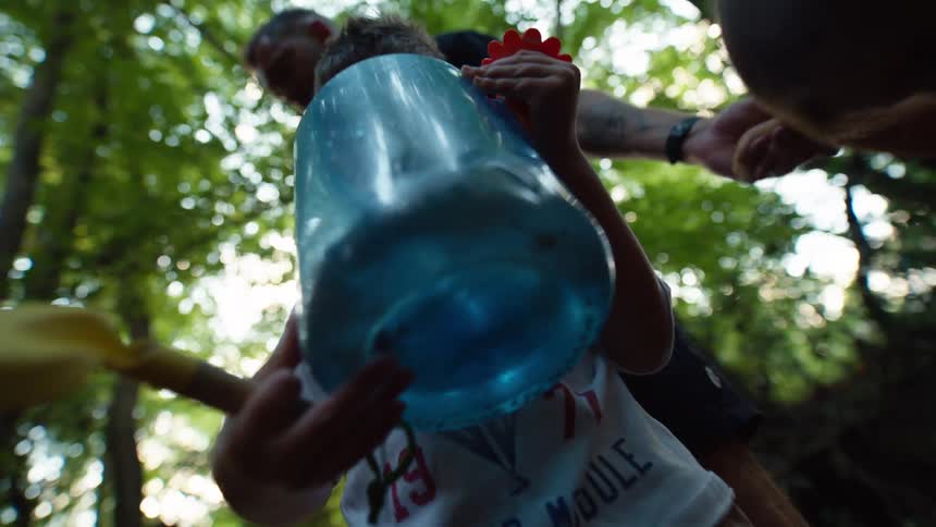 A family is seen from a low angle with one child holding a large blue bottle of water and a red shovel while his family stands around