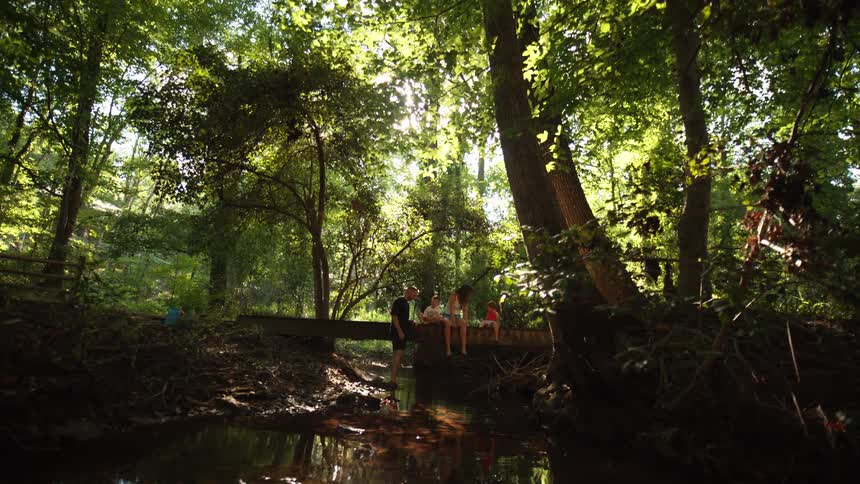 A small family sits and dangles their feet over the side of a wooden bridge looking at the water in a small calm stream surrounded by tall green trees