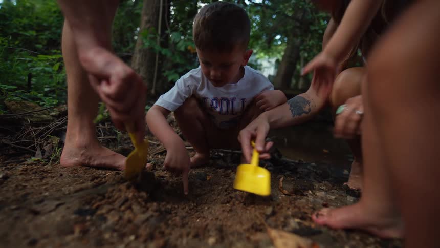 Male toddler squatting and playfully digging on the soil together with a female toddler and a couple of adults outdoors during day time
