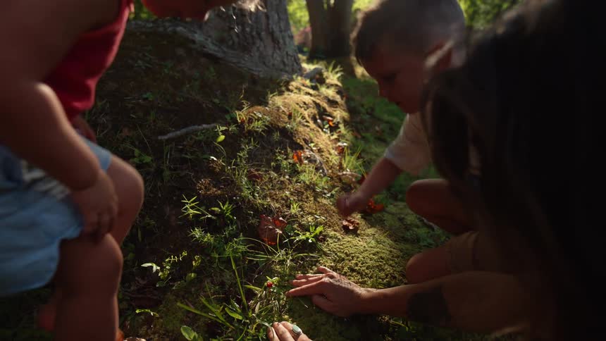 A woman looks through the moss on a tree root showing her two children how to look for mushrooms