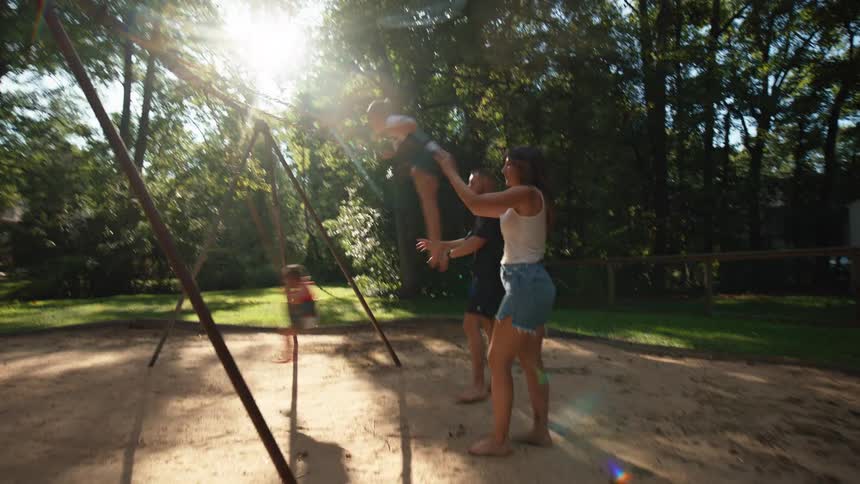 Parents pushing their children on a swing set in an outdoor playground setting on a sunny day