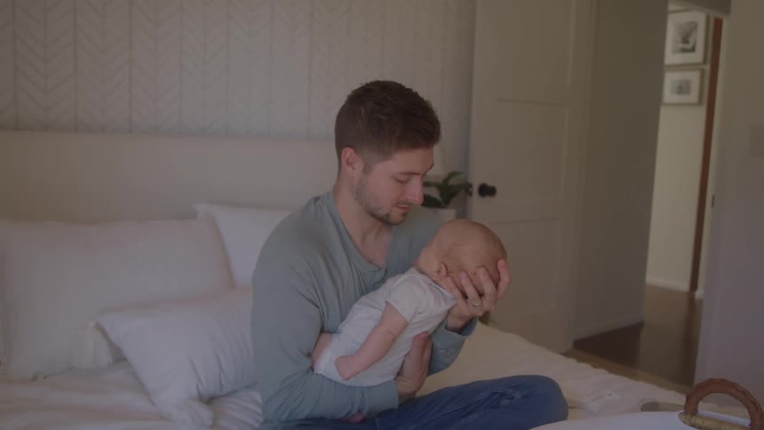 A man sits on a bed with a white headboard and holds a small swaddled baby in his arms looking down at the baby with a smile
