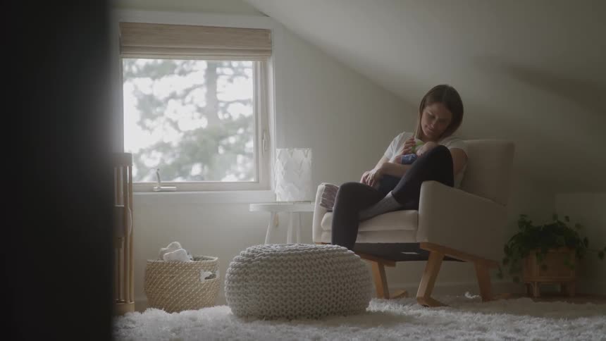 A person sits in a rocking chair and holds a baby in a room with a window and plants