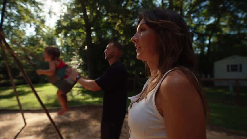 A mother and father push two children on swings on a sandy playground with a house and trees in the background