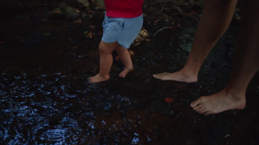A small child wearing a red tank top and blue shorts holds a yellow shovel and is guided by a persons hand as they walk barefoot in a shallow stream