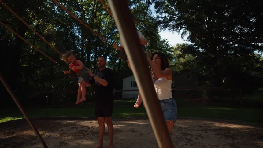 A mother and father stand side by side on a sandy playground and push their two young children on swings with a house and trees in the background