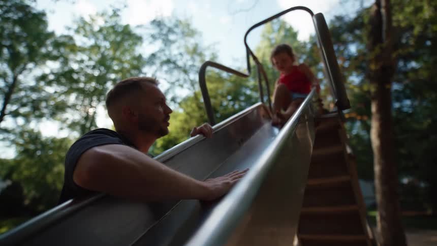 Father watches daughter as she slides down a slide in an outdoor play area during daytime