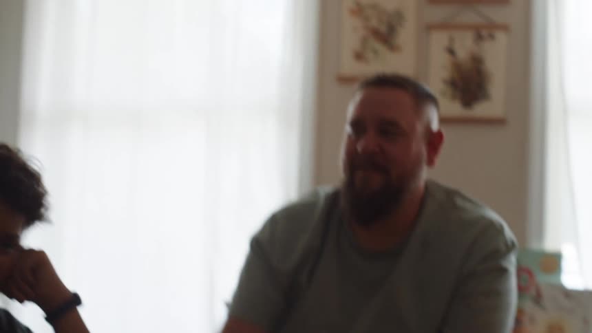 A medium shot captures a man with a beard and a boy who are out of focus in the foreground another persons hand is raised in the foreground as they speak to the man