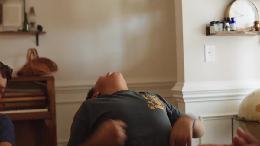 A boy sits at a table and laughs while reacting to a person offcamera