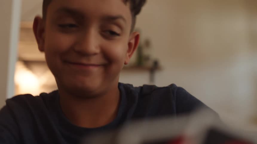 A boy with a short haircut and a dark shirt looks at a deck of cards then closes his eyes and smiles before looking back up with a big smile and a wide grin