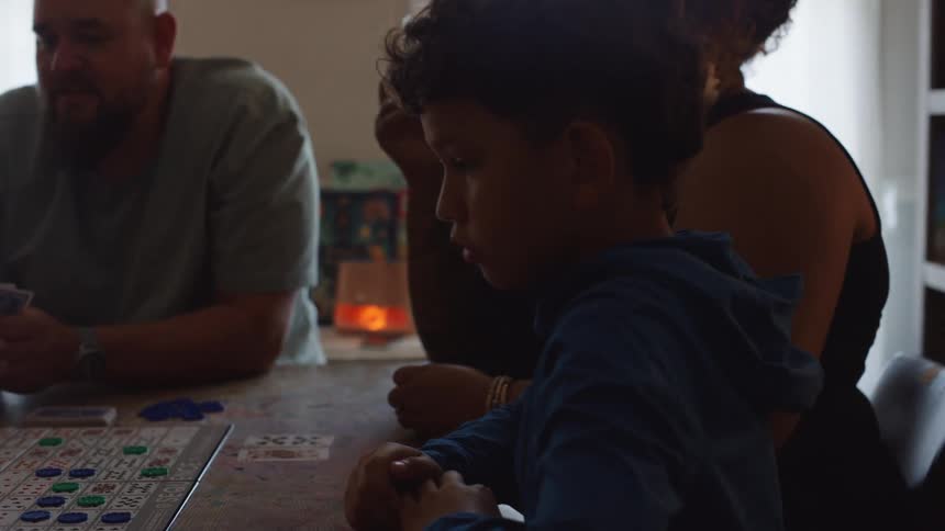 Child watches a board closely while adults play cards around the table