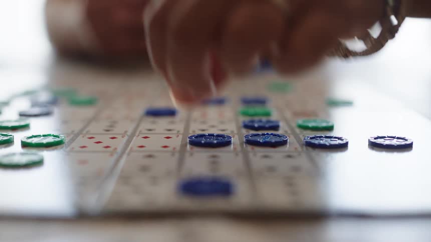 A person with rings and bracelets on their right hand places a blue piece onto a game board with numbers and red dots next to several other green and blue pieces
