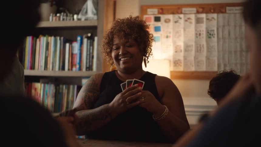 A group of friends playing cards on a library setting the camera focusing on a black woman with earrings and a bracelet