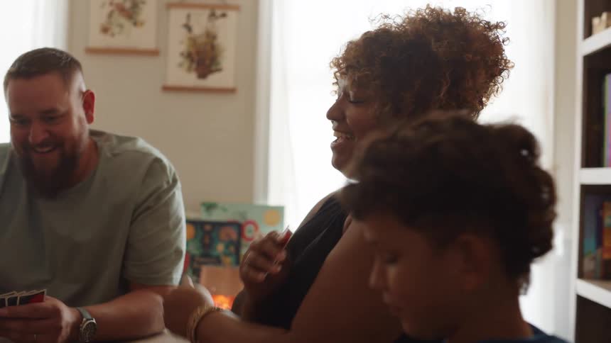 A diverse family of a mother father and three children sit around a table and play a card game