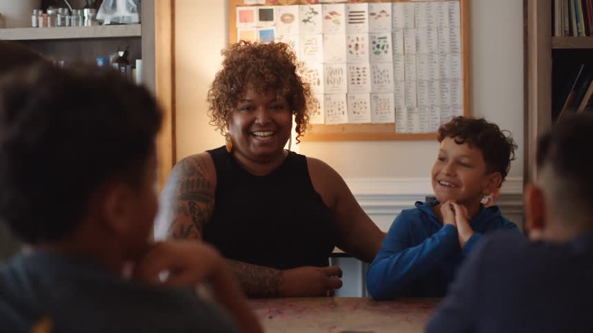 A happy teacher with curly hair and visible arm tattoos sits at a table with smiling students in a classroom with bookshelves and charts on the wall