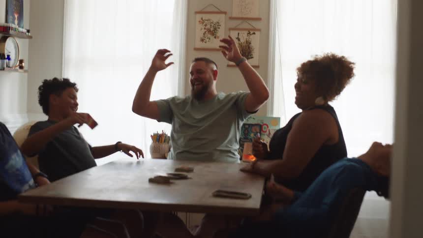 A diverse family of a mother father and three children sit around a table and play a card game with the father and a son laughing and giving each other a high five