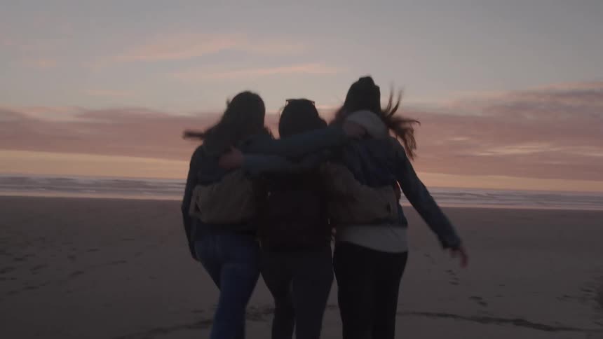 The females skip along a beach with their arms around each other